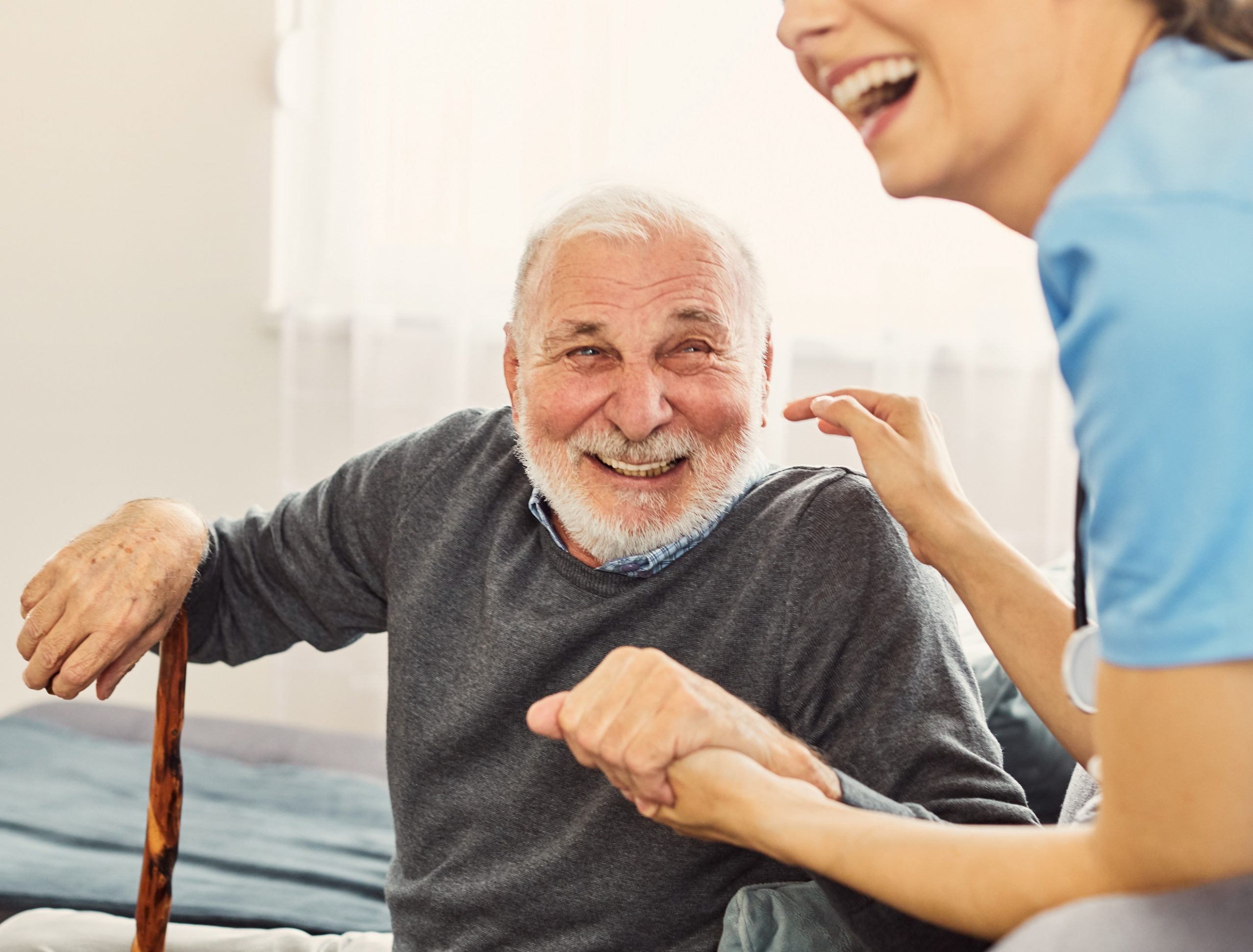 An elderly man with a walking stick and a young caregiver are laughing