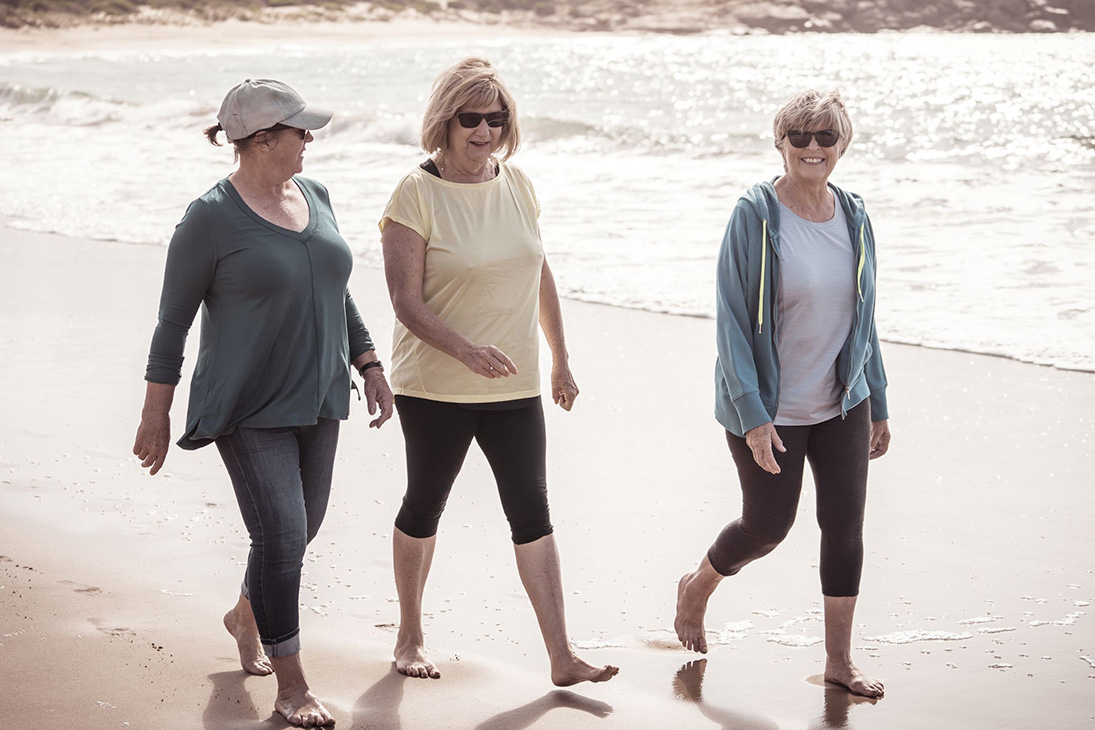 Three women are walking barefoot along the beach and talking.